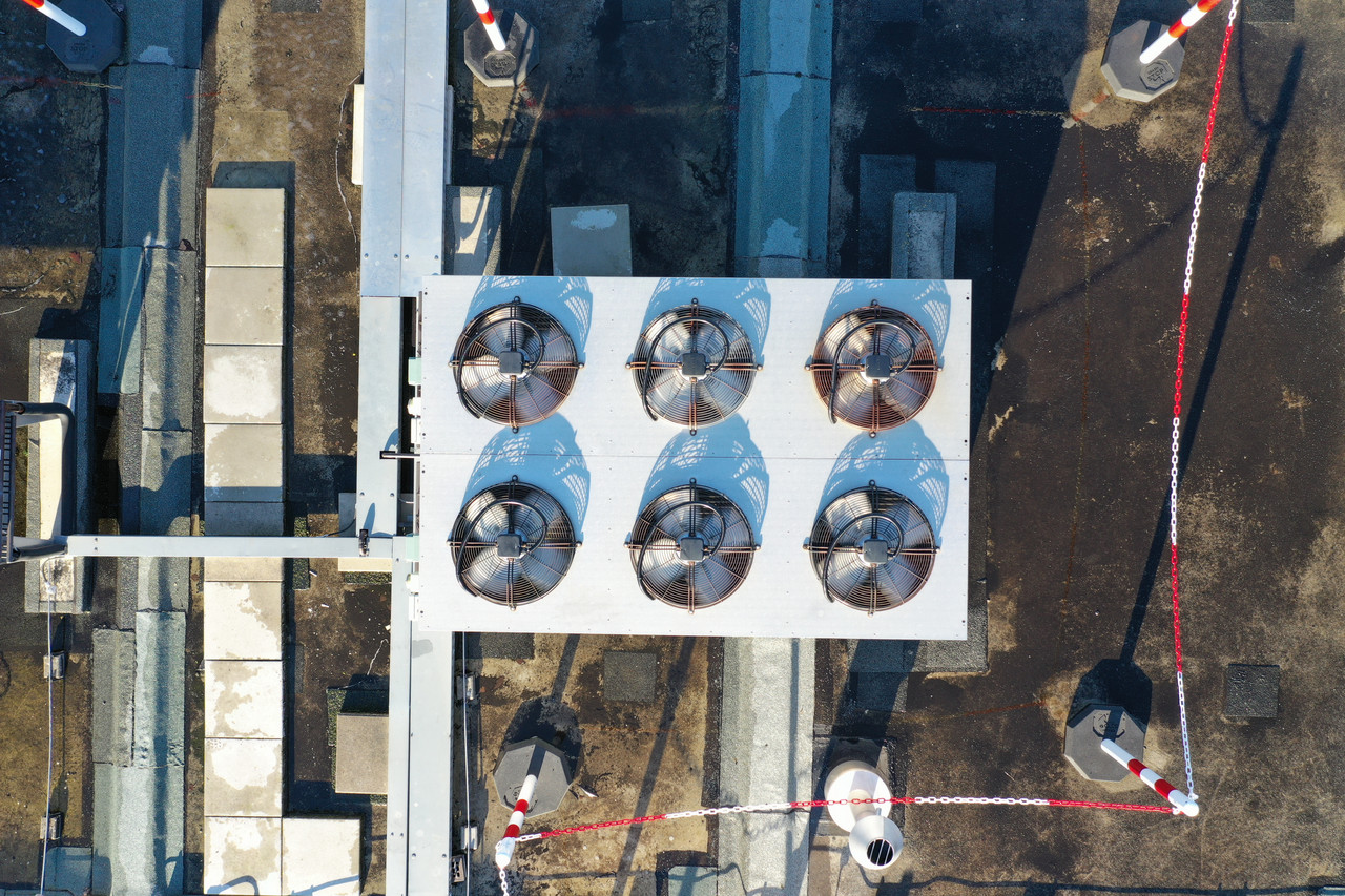 Luftaufnahme von sechs großen Lüftern oder Ventilatoren auf dem Dach eines Gebäudes, vermutlich Teil eines Kühlsystems für ein Rechenzentrum. Die Lüfter sind in zwei Reihen zu je drei Einheiten angeordnet und befinden sich innerhalb einer markierten Fläche, die mit rot-weißen Absperrbändern gesichert ist. Die Umgebung zeigt verschiedene Dachstrukturen und Installationen, die auf eine technische Umgebung hindeuten.