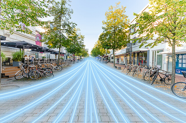 Foto einer Einkaufsstraße mit Cafés, Geschäften und Bäumen an den Seiten, in die eine digitale Illustration von leuchtenden blauen Linien eingefügt wurde, die in die Ferne verlaufen. Die Linien symbolisieren möglicherweise eine schnelle Datenübertragung oder ein Highspeed-Netzwerk, das durch die Straße fließt. Fahrräder sind entlang des Gehwegs abgestellt, und die Atmosphäre wirkt freundlich und modern.