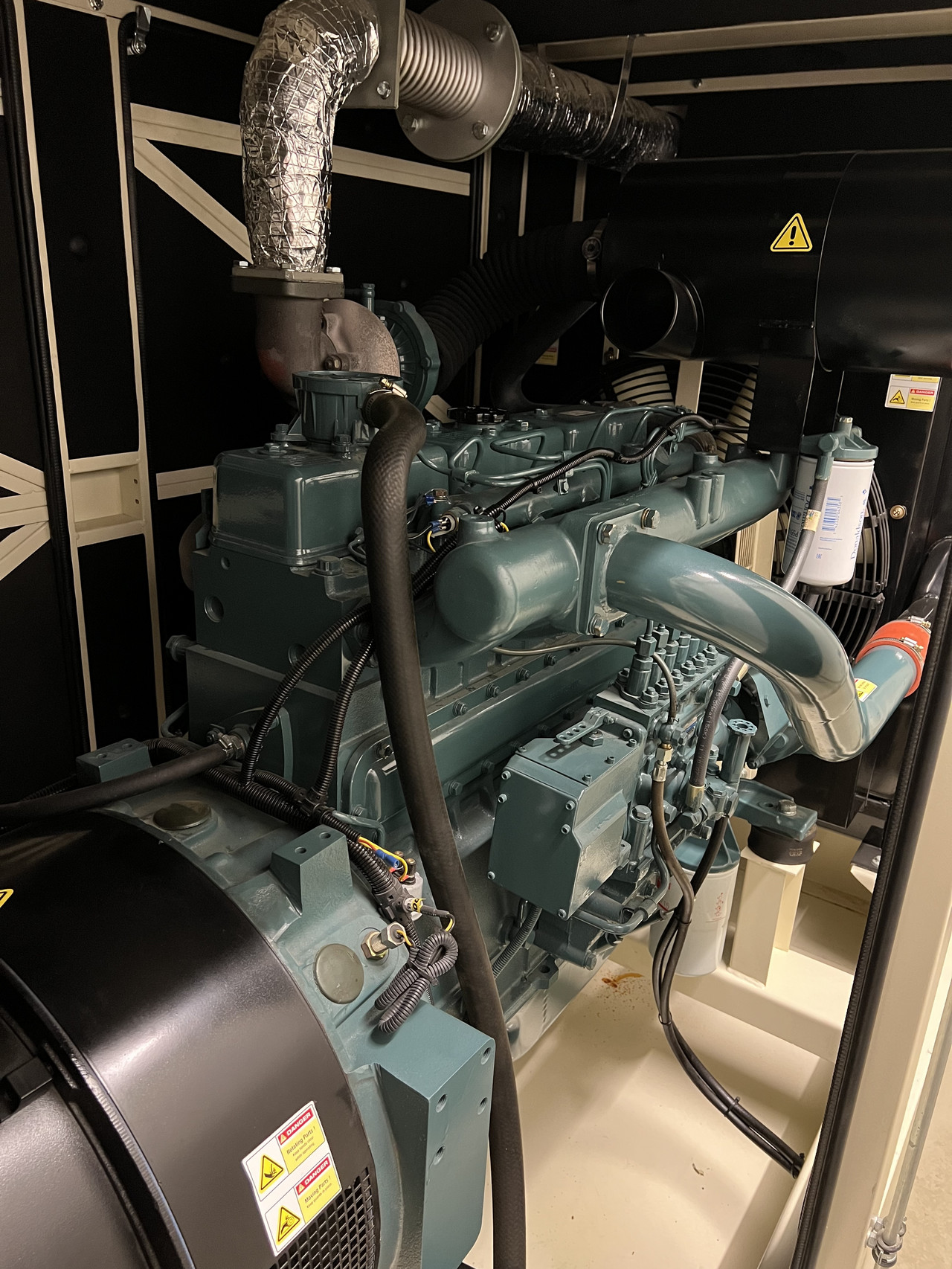 Detailaufnahme eines großen Dieselgenerators oder Notstromaggregats in einem Technikraum. Das Gerät ist in einem dunklen Gehäuse installiert und zeigt komplexe Rohrleitungen, Kabelverbindungen und eine isolierte Abgasleitung. Der Generator ist in einem metallischen Grün und Schwarz gehalten, mit mehreren Warnhinweisen, die auf die Sicherheitsvorkehrungen hinweisen. Solche Generatoren dienen oft zur Sicherstellung der Stromversorgung in Rechenzentren oder anderen kritischen Infrastrukturen bei einem Stromausfall.