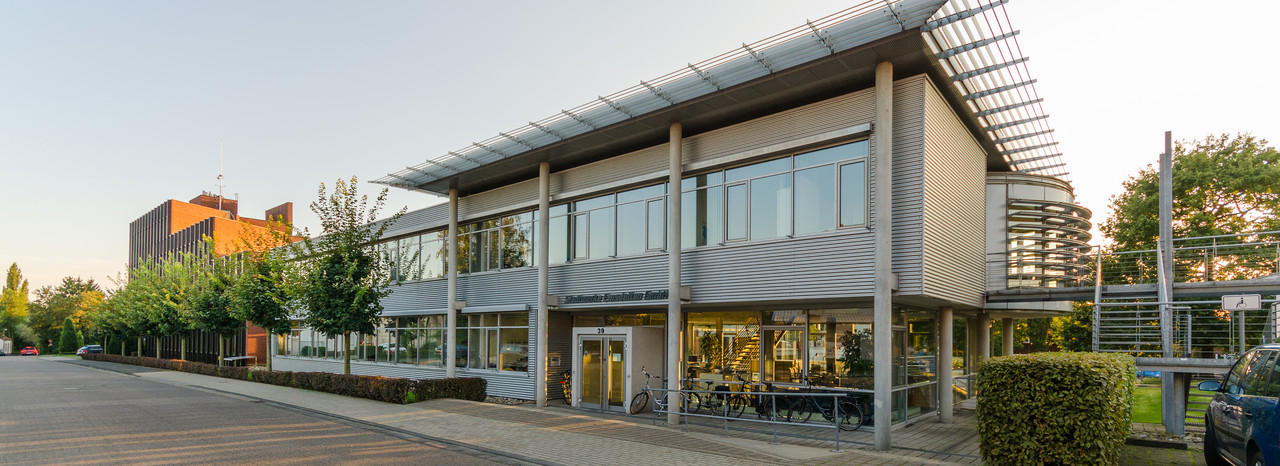 Außenansicht eines modernen Bürogebäudes im Abendlicht, mit großen Glasfenstern und einer metallischen Fassade. Das Gebäude hat zwei Stockwerke und ist von Bäumen und Grünanlagen umgeben. Auf dem Gehweg davor sind Fahrräder abgestellt. Links im Hintergrund ist ein weiteres, größeres Gebäude zu sehen. Die Architektur vermittelt eine offene und einladende Atmosphäre.
